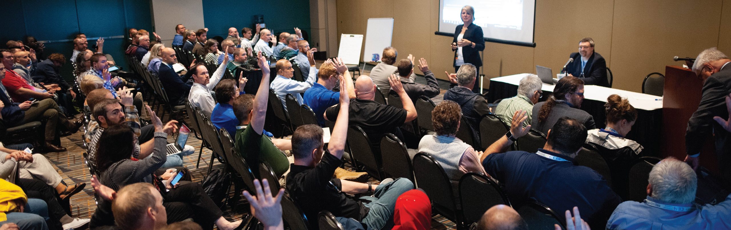 Image from the CEN Conference of a group of attendees sitting in chairs raising their hands and a woman speaking and giving a presentation